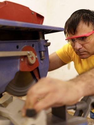 Carpenter cutting a piece of wood for furniture in his woodwork workshop, using a circular saw, and wearing safety goggles and earmuffs.